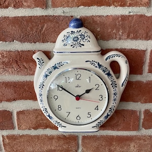 Op de afbeelding: Een crèmekleurige keramische wandklok in de vorm van een theepot, voorzien van blauwe bloemmotieven, een zilverkleurige rand, zwarte uren- en minutenwijzers en een rode secondewijzer. De wijzerplaat toont de merknaam Pecer en het woord quartz.