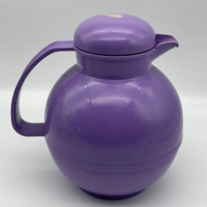 Könnte beinhalten: Eine kugelförmige, violette Thermokanne mit passendem Deckel, einem geschwungenen Griff an der Seite und einem kleinen Ausgießer. Die Oberfläche des Kunststoffbehälters ist glatt und glänzend und weist ein kleines, rundes Etikett auf dem Deckel auf.