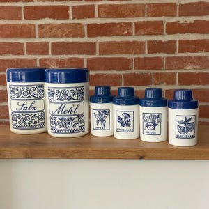 Op de afbeelding: Een set van zes witte keramische keukenpotten met donkerblauwe deksels en decoratieve blauwe patronen. De twee grotere potten zijn gelabeld met Salz en Mehl, terwijl de vier kleinere potten illustraties en labels hebben voor Paprika, Kümmel, Pfeffer en Muskat.
