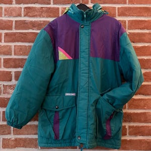 Könnte beinhalten: Eine Vintage-Winterjacke in Teal, Lila und Grün mit Kapuze. Die Jacke hat ein Colorblock-Design mit einem geometrischen Akzent auf der oberen linken Brust. Sie hat zwei Vordertaschen und einen Kordelzug am unteren Saum. Die Jacke hängt an einem schwarzen Kleiderbügel.