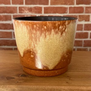 Può includere: Un vaso di ceramica con smalto marrone e beige. Il vaso ha un bordo marrone scuro e una base a sottovaso abbinata. Lo smalto ha un effetto gocciolante, creando una trama unica. Circa 15 cm di altezza.