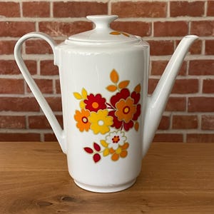 Op de afbeelding: Witte keramische koffiepot met een deksel en een lange tuit. De pot is versierd met een bloemenpatroon in tinten rood, oranje en geel. De handgreep is gebogen en de pot staat op een houten oppervlak.