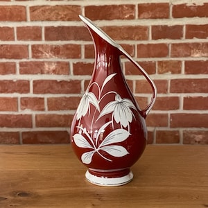 Op de afbeelding: Een dieprode keramische kan met een wit bloemmotief van puntige bloemblaadjes en bladeren. De kan heeft een gebogen handvat, een taps toelopende hals, een brede ronde buik en een witte voetrand. Het ontwerp is glad met een glanzende afwerking.