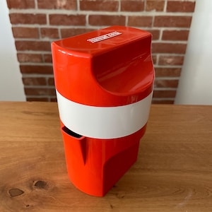 Op de afbeelding: Een feloranje en witte citruspers van kunststof met een verticaal, cilindrisch ontwerp. Het bovenste gedeelte is voorzien van een gevormde handgreep en de in reliëf aangebrachte tekst "Koziol Plastic" op het deksel. Een brede witte band omcirkelt het midden.