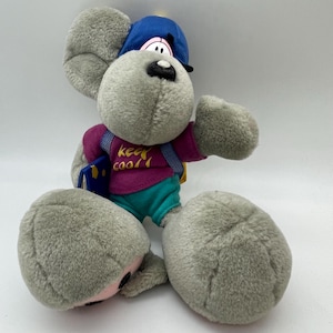 Könnte beinhalten: Ein Plüschtier, das eine Maus darstellt, die eine blaue Baseballkappe, ein lila Hemd mit dem Text "keep cool" und türkisfarbene Shorts trägt. Die Maus hat graues Fell, große Ohren und einen Rucksack. Sie hält ein kleines blaues Buch.