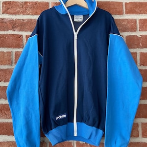 Könnte beinhalten: Eine marineblaue und hellblaue Vintage-Trainingsjacke mit Reißverschluss. Die Jacke hat hellblaue Ärmel und einen weißen Reißverschluss. Der Markenname "Sporett" ist sichtbar. Sie hängt an einem Holzbügel vor einer Backsteinmauer.