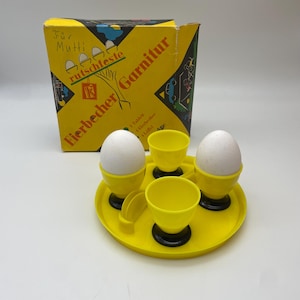 Può includere: Un set da portata per uova giallo vintage con due uova in portauova gialli, due tazze vuote e due cucchiai su un vassoio giallo. Il set include una scatola gialla con testo e illustrazioni in tedesco.