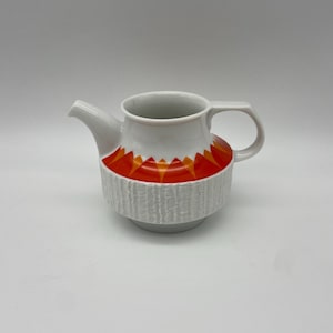 Op de afbeelding: Witte keramische theepot met een getextureerde, geribbelde onderkant en een levendig oranje en geel geometrisch ontwerp. Voorzien van een tuit en een gebogen handvat. Een decoratief stuk voor het serveren van thee.
