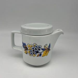 May include: A white ceramic teapot with a cylindrical body, a flat lid, and a side handle. The exterior features a decorative floral pattern in shades of yellow, blue, and brown, wrapping around the center of the vessel with a small spout.