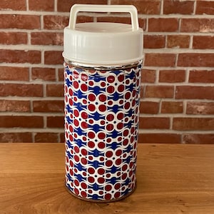 Könnte beinhalten: Eine isolierte Vintage-Thermoskanne mit weißem Griff und Deckel. Der zylindrische Körper weist ein sich wiederholendes Muster aus roten und weißen Kreisen auf blauem Hintergrund auf. Die Thermoskanne ist für heiße oder kalte Getränke konzipiert.