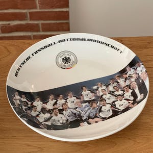 Op de afbeelding: Een witte keramische kom met een afbeelding van de spelers van het Duitse nationale voetbalelftal in hun witte shirts. De tekst DEUTSCHE FUSSBALL-NATIONALMANNSCHAFT staat langs de rand en het officiële embleem staat in het midden.