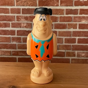 Könnte beinhalten: Eine Vintage-Figur von Fred Feuerstein. Die Figur ist beige mit einer schwarzen Haube und einer orangefarbenen Tunika mit schwarzen Dreiecken und einer türkisfarbenen Krawatte. Die Figur steht auf einem beigen Sockel.