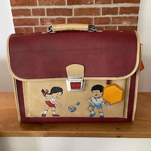 Könnte beinhalten: Vintage Schulranzen in Burgunderrot und Beige mit Tennis-Illustration. Der Ranzen hat einen Metallverschluss, einen beigefarbenen Griff und ein sechseckiges Anhängerschild. Die Illustration zeigt zwei Kinder beim Tennisspielen.