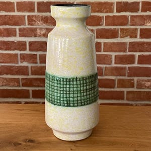 Può includere: Un vaso in ceramica con corpo cilindrico e bordo svasato. La superficie presenta uno smalto screziato color crema e giallo, accentuato da un'ampia fascia orizzontale testurizzata con motivi a griglia verde scuro che circonda il centro.