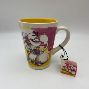 Könnte beinhalten: Weißer Keramikbecher mit gelbem Innenraum und einer Cartoon-Maus-Illustration. Die Maus wird beim Malen mit einer Farbpalette dargestellt. Ein kleines Etikett mit passender Illustration hängt am Griff.