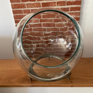Könnte beinhalten: Eine große, kugelförmige Schale aus klarem Glas mit einer breiten, schrägen Öffnung an der Oberseite. Die Schale ruht auf einem kreisförmigen, silberfarbenen Metallständer mit drei kurzen, stabilen Beinen, die dem Glasgefäß Halt geben.