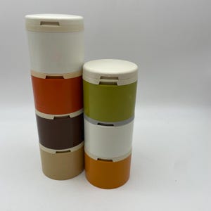 Op de afbeelding: Gestapelde, cilindrische containers in verschillende kleuren. De containers hebben witte deksels en zijn in de tinten wit, oranje, bruin, groen en beige. Ze zijn waarschijnlijk gemaakt van plastic en bedoeld voor opslag.