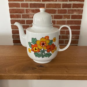Könnte beinhalten: Weiße Keramik-Teekanne mit Deckel und Henkel. Die Teekanne ist mit einem Blumendesign in Orange, Gelb und Grün verziert. Der Deckel hat einen orangefarbenen Knopf. Die Teekanne steht auf einer Holzoberfläche.