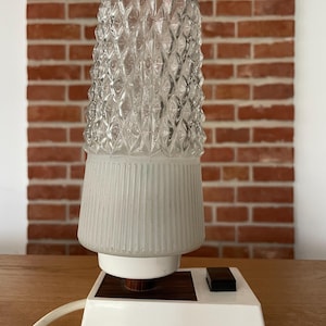 Op de afbeelding: Een vintage tafellamp met een heldere, getextureerde glazen kap en een witte voet. De kap heeft een diamantpatroon en de voet heeft een wit, bruin en zwart ontwerp met een schakelaar. De lamp is aangesloten.