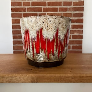 Könnte beinhalten: Ein Keramik-Pflanzgefäß mit einzigartigem Design. Die Oberseite ist mit Braun und Creme gesprenkelt und geht in ein leuchtend rotes Muster über. Der Sockel ist dunkelbraun und erzeugt einen auffälligen Kontrast. Dieses Pflanzgefäß ist ideal für Zimmerpflanzen.