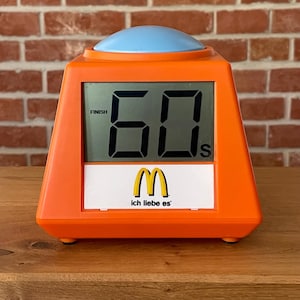 Könnte beinhalten: Orangener digitaler Timer mit hellblauer Taste. Das Display zeigt "60 S" und "FINISH". Das McDonald's-Logo und der Schriftzug "ich liebe es" sind auf der Vorderseite aufgedruckt. Ein Retro-Küchengerät.