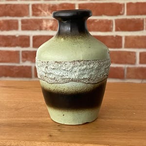 Può includere: Un vaso in ceramica con corpo bulboso, collo stretto e bordo svasato. La superficie presenta una sfumatura di toni verde chiaro e marrone scuro, accentuata da un'ampia fascia testurizzata color bianco sporco che circonda il centro del vaso.