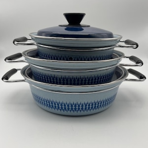 Op de afbeelding: Een stapel van vier blauw-witte emaille potten en pannen. De set bevat een donkerblauw deksel met een zwarte handgreep. De potten hebben zilveren randen en zwarte handgrepen. De buitenkant van de potten heeft een blauw-wit geometrisch patroon.