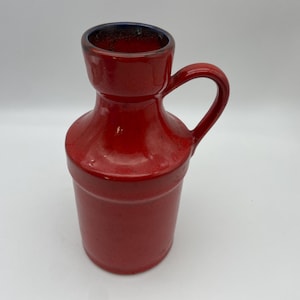 Può includere: Una brocca in ceramica rosso brillante con corpo cilindrico, collo svasato e un singolo manico curvo sul lato. L'interno del bordo presenta uno smalto blu scuro, mentre la superficie esterna ha una finitura rossa liscia e lucida.