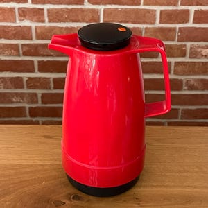 Può includere: Una caraffa isolante rossa con coperchio e base neri. La caraffa ha un manico e un beccuccio per versare. Il coperchio ha un piccolo punto arancione. La caraffa è appoggiata su una superficie di legno.