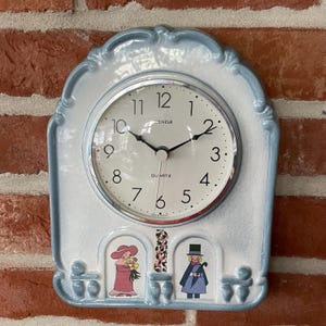 Op de afbeelding: Een lichtblauwe en witte keramische wandklok met een afgeronde bovenkant. De wijzerplaat heeft zwarte cijfers, zwarte wijzers en de tekst Kienzle Quartz. Onder de wijzerplaat staan twee kleine illustraties van figuren en bloemen.