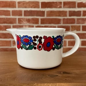 Op de afbeelding: Witte keramische creamer met een bloemenontwerp in rood, blauw en groen. De creamer heeft een handvat en een schenktuit. Het bloemenpatroon wordt herhaald rond de body van de creamer.