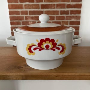 May include: White ceramic serving dish with a lid and handles. The lid has a brown band and a white knob. The dish is decorated with a red and yellow floral pattern. A vintage kitchenware item.