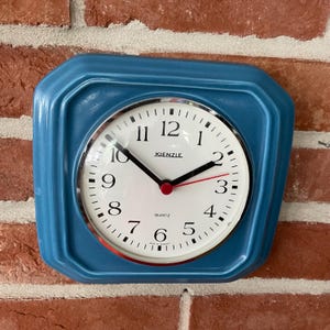 Könnte beinhalten: Eine quadratische blaue Wanduhr mit weißem Zifferblatt, schwarzen Ziffern sowie schwarzen Stunden- und Minutenzeigern. Ein dünner roter Sekundenzeiger ist mittig angebracht. Der Markenname Kienzle steht in Schwarz über der Mitte, darunter Quartz.