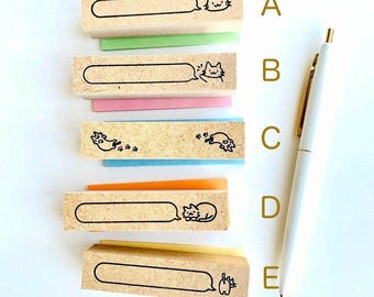 May include: Five wooden stamps with cat illustrations and speech bubbles, each on a different colored sticky note. A white pen with gold accents is positioned vertically on the right side of the image. The stamps feature various cat designs.