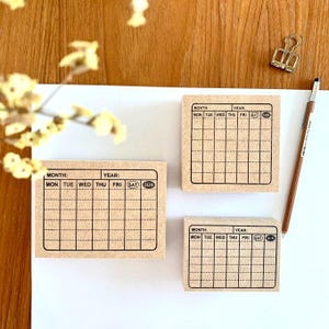 May include: Three rectangular brown stamps with a weekly calendar layout. Each stamp has the days of the week printed in black, with space for the month and year. A pencil and a gold paper clip are also visible.