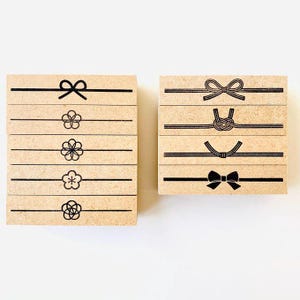 Op de afbeelding: Twee houten blokken met zwarte lijntekeningen. Eén blok heeft een strik, een bloem en andere bloemen en geometrische patronen. Het andere blok heeft een strik, een knoop en andere strikachtige ontwerpen. De blokken staan op een witte achtergrond.