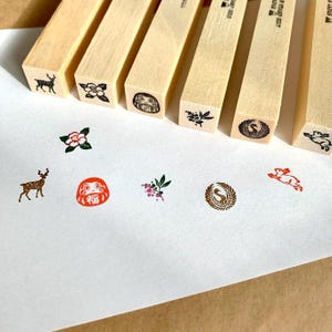 Puede incluir: Sellos de madera con varios diseños de inspiración japonesa, incluyendo un ciervo, una flor, una muñeca Daruma y un pájaro dentro de un círculo. Los sellos están dispuestos sobre una superficie blanca, con sus imágenes estampadas correspondientes debajo.