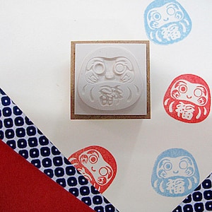 May include: A set of five rubber stamps featuring a traditional Japanese Daruma doll design. The stamps are in various colors, including red, blue, and white. The Daruma dolls have a smiling face and are holding a lucky charm. The stamps are mounted on wooden blocks.