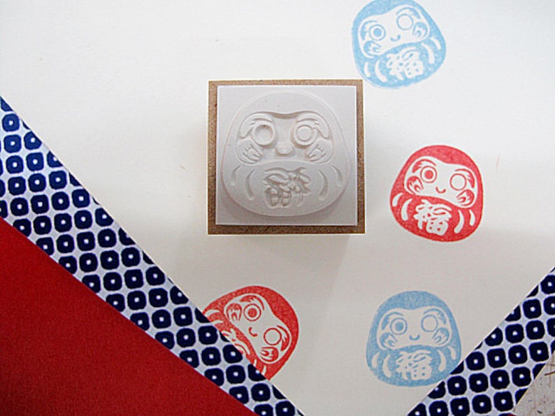 May include: A set of five rubber stamps featuring a traditional Japanese Daruma doll design. The stamps are in various colors, including red, blue, and white. The Daruma dolls have a smiling face and are holding a lucky charm. The stamps are mounted on wooden blocks.