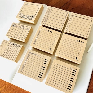 May include: A selection of brown paper memo pads with different designs. Some pads have lined pages, checklists, and calendars. Others feature illustrations of cats, birds, and musical notes. The pads are arranged on a white surface.