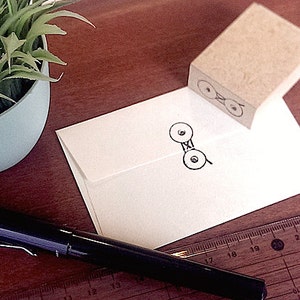 May include: A still-life composition featuring stationery items. A white envelope with a black stamp, a black pen, and a wooden ruler are arranged on a wooden surface. A small potted plant is in the background. The stamp design is repeated on the envelope.