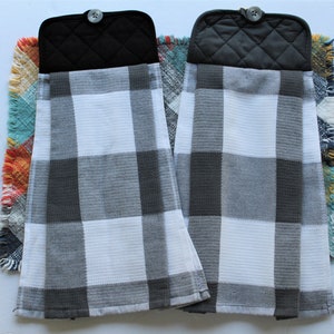 May include: Two black and white checkered kitchen towels with black quilted pot holders attached. The towels have a frayed edge and are hanging on a colorful woven rug.