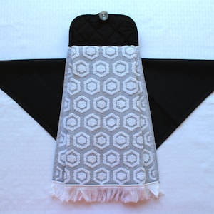 May include: A gray and white kitchen towel with a geometric pattern and a black quilted potholder with a button. The towel has a fringe border.
