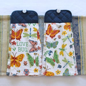 May include: Two hanging kitchen towels with a white background, featuring colorful butterflies, dragonflies, and flowers. The towels have a navy blue quilted top with a white button. One towel has the text "LOVE BUG".
