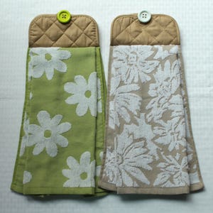 May include: Two hanging kitchen towels. One is light green with white daisy patterns, the other is beige with white floral patterns. Both have a quilted brown top with a button for hanging.