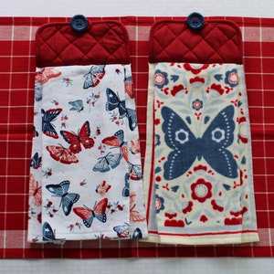 May include: Two red quilted pot holders with blue buttons, each holding a white tea towel with a blue and red butterfly pattern. The tea towels are folded and hanging from the pot holders.