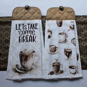 May include: Two hanging kitchen towels. One towel has the text "LET'S TAKE A COFFEE BREAK" and a coffee cup graphic. The other towel has a pattern of coffee cups. Both towels have a quilted top and a button.
