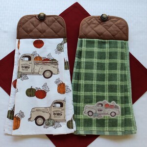 May include: Two kitchen towels with a fall theme. One towel is white with a brown truck carrying pumpkins and the text "Pumpkin Patch". The other towel is green with a plaid pattern and a brown truck carrying pumpkins. Both towels have brown quilted fabric on the top with a button.