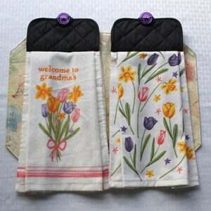 May include: Two white tea towels with black quilted pot holders. One towel has a floral design with the text "welcome to grandma's". The other towel has a floral design with yellow daffodils and purple tulips.