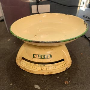 May include: A vintage cream-coloured kitchen scale with a green-trimmed bowl. The scale features a curved face with measurements in pounds and the brand name "Salter" in a green rectangle. Made in England.